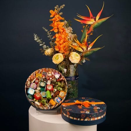 Tropical floral arrangement featuring orange orchids, bird of paradise flowers, and cream roses with greenery in an elegant vase.