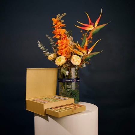 Tropical floral arrangement featuring orange orchids, bird of paradise flowers, and cream roses with greenery in an elegant vase.