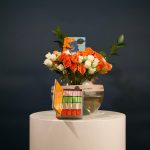 Rounded floral centerpiece with orange, blue, and ivory flowers arranged in a bowl-shaped vase with lush greenery.