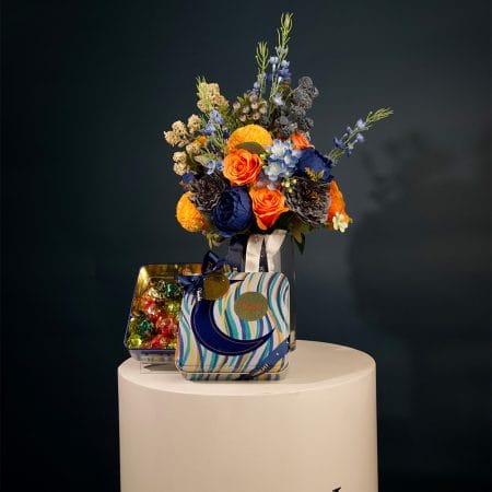 Colorful floral arrangement with golden yellow pom-pom flowers, orange roses, and deep blue blooms arranged in a clear glass vase with decorative ribbons.