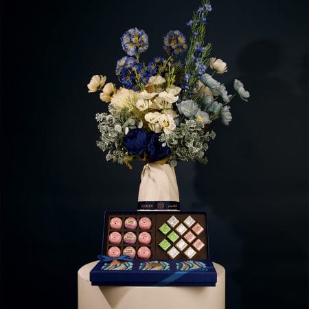 Elegant floral arrangement featuring deep blue, ivory, and soft blue blossoms with lush greenery, arranged in a sculptural ceramic vase.