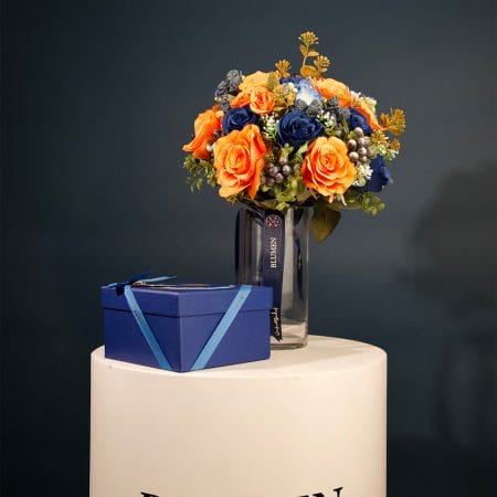 Floral arrangement with bright amber-orange roses, deep blue flowers, and greenery arranged in a clear glass vase with decorative ribbon.