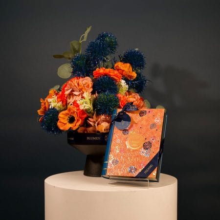 Elegant floral arrangement featuring deep blue spherical flowers and coral-toned blossoms in a modern black pedestal vase.