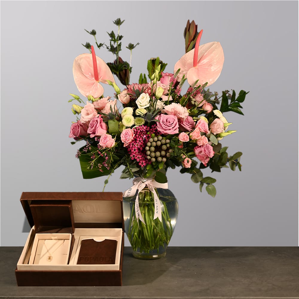 A lush floral arrangement in a clear glass vase featuring pink roses, anthuriums, and mixed greenery with a soft blush color palette.