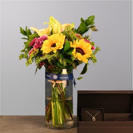 A vibrant bouquet with sunflowers, lilies, and greenery in a clear glass vase, paired with an elegant open jewelry box displaying a necklace.