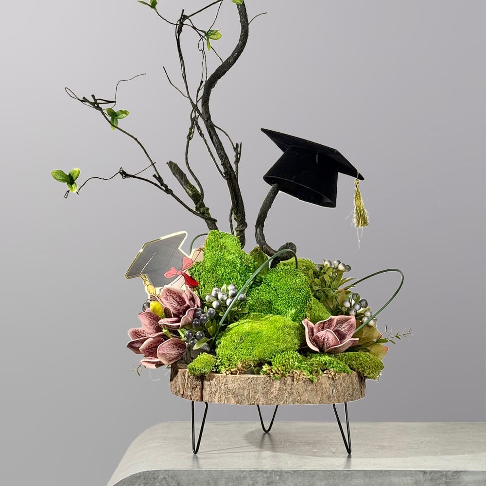 A modern botanical arrangement featuring preserved green moss, sculptural branches, floral accents, and graduation-themed decorations on a wooden display stand.