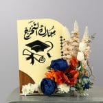 A graduation-themed floral arrangement featuring blue and orange flowers, white tulips, and a congratulatory sign with a graduation cap illustration.