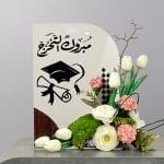 A graduation-themed floral arrangement with white tulips, white blooms, pink carnations, green textured flowers, and a congratulatory graduation display sign.