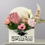 A graduation-themed floral arrangement in a decorative envelope-style box featuring pink protea, white tulips, soft pink flowers, and lush greenery.