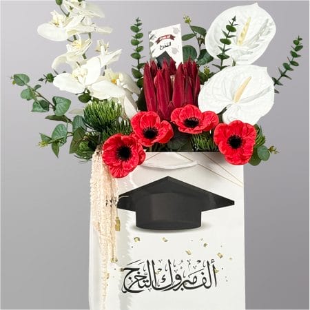 A graduation-themed floral arrangement in a decorative gift bag featuring white orchids, white anthuriums, red blooms, protea flowers, and lush greenery.