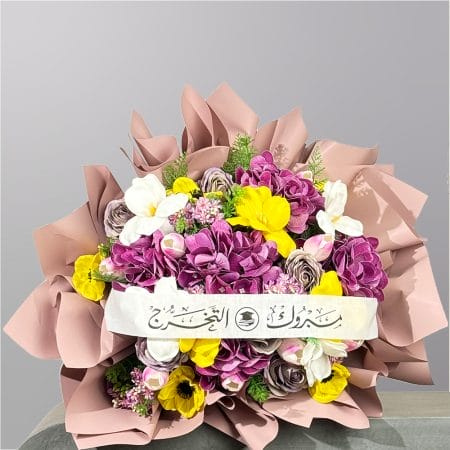 A luxurious graduation bouquet wrapped in blush paper featuring purple hydrangeas, yellow flowers, white orchids, and soft pastel roses with a congratulatory ribbon.