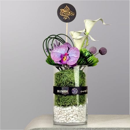 A contemporary floral arrangement featuring a purple orchid, white calla lilies, decorative greenery, and moss displayed in a clear cylindrical glass vase with white stones.