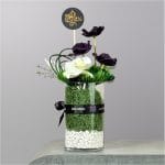 A modern floral arrangement featuring deep purple flowers, a white orchid bloom, decorative greenery, moss, and white stones displayed in a cylindrical glass vase.