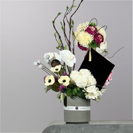 A modern graduation floral arrangement featuring white roses, anthuriums, decorative branches, purple accents, and a black graduation cap centerpiece in a textured vase.