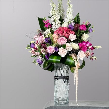 A luxurious floral arrangement featuring pink roses, white blooms, purple flowers, and vibrant greenery arranged in a tall crystal-style glass vase.