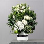 A luxurious white floral arrangement featuring hydrangeas, roses, lisianthus, green button chrysanthemums, and white gerberas arranged in a modern white vase with flowing greenery accents.