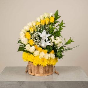 A luxurious floral arrangement featuring white roses, yellow roses, white lilies, and seasonal blooms arranged in a rustic wooden container.