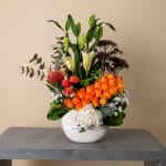 A luxurious floral arrangement featuring orange roses, white lilies, tulips, hydrangeas, and exotic foliage arranged in a contemporary white ceramic bowl.