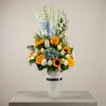 A luxury floral arrangement featuring sunflowers, yellow gerberas, blue hydrangeas, white roses, and delphiniums arranged in a tall white vase.