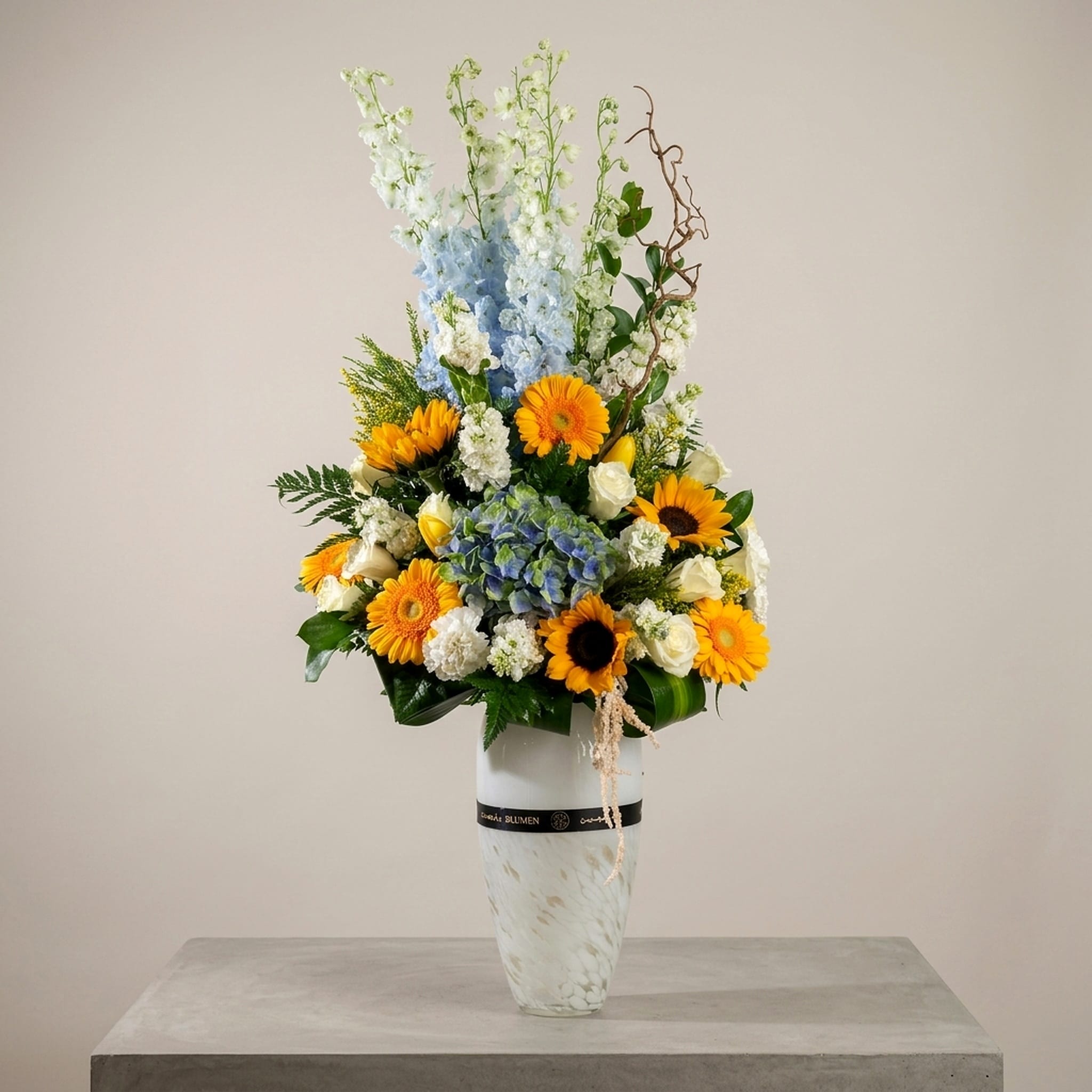 A luxury floral arrangement featuring sunflowers, yellow gerberas, blue hydrangeas, white roses, and delphiniums arranged in a tall white vase.
