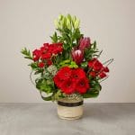 A luxury floral arrangement with red spray roses, red gerbera daisies, king proteas, lily buds, and decorative greenery in an elegant ceramic container.