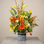 A luxury floral arrangement featuring yellow calla lilies, orange orchids, orange gerbera, tulips, and lush greenery in a modern stone vase.
