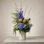 The Celestial Tide floral arrangement featuring blue hydrangea, blue delphinium, yellow pincushion protea, ivory anthuriums, and cascading greenery in a textured white vase.