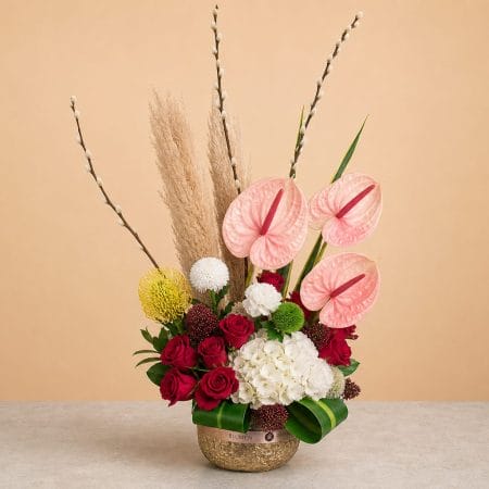 Luxury floral arrangement featuring blush pink anthuriums, red roses, white hydrangea, pampas grass, pussy willow branches, and seasonal blooms in a textured golden vase.