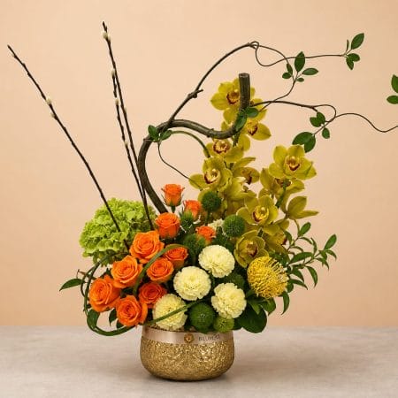 Luxury floral arrangement featuring green cymbidium orchids, orange roses, white carnations, green hydrangea, pincushion protea, decorative branches, and lush foliage in a golden vase.