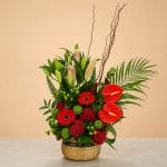 Luxury floral arrangement featuring red anthuriums, red gerbera daisies, red cockscomb blooms, pink lilies, green button chrysanthemums, tropical foliage, and decorative branches in a gold vase.