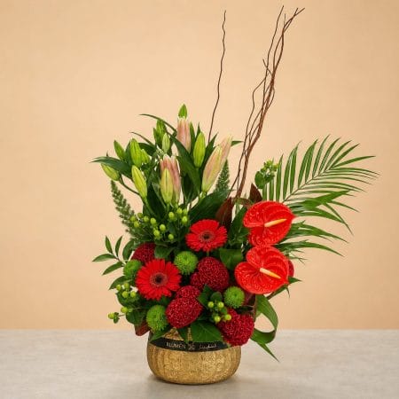 Luxury floral arrangement featuring red anthuriums, red gerbera daisies, red cockscomb blooms, pink lilies, green button chrysanthemums, tropical foliage, and decorative branches in a gold vase.