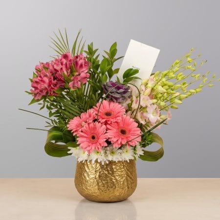A luxurious floral arrangement featuring vibrant pink gerberas, blush roses, orchids, and premium seasonal flowers, beautifully arranged in an elegant gold vase.