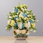 A sophisticated floral arrangement featuring elegant white lilies, soft blue and white blooms, and lush seasonal greenery, beautifully arranged in a modern decorative vase.