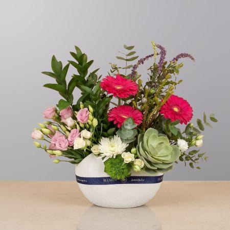 A contemporary floral arrangement featuring vibrant gerberas, delicate roses, lush greenery, succulents, and premium seasonal flowers, beautifully arranged in a modern ceramic bowl.
