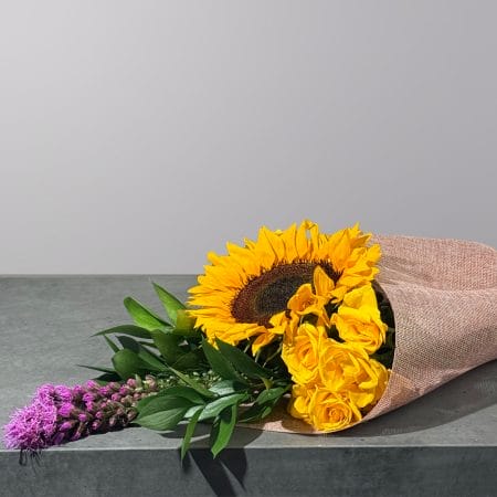 Rustic bouquet featuring a vibrant sunflower, golden yellow roses, purple liatris, and fresh greenery wrapped in natural fabric.