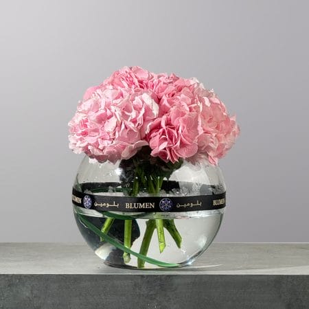 The Petal Sonata featuring fresh blush pink hydrangeas artistically arranged in a clear round glass bowl with water.