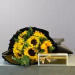 A lush bouquet of bright yellow sunflowers and fresh green foliage wrapped in elegant black paper.