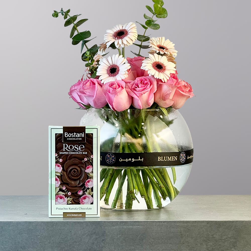 A rounded glass vase filled with soft pink roses and white and peach gerbera daisies, accented with fresh eucalyptus greenery.
