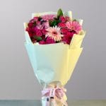 A vibrant hand-tied bouquet of pink gerbera daisies and roses in various shades of pink and fuchsia, wrapped in soft pastel paper with a delicate ribbon.
