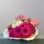 A vibrant bouquet of fuchsia gerbera daisies, hot pink spray roses, and creamy white roses wrapped in elegant coral-pink paper.