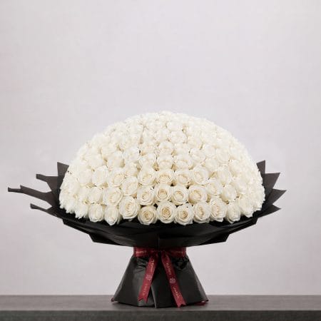 A large bouquet of elegant white roses arranged in a full, rounded shape, wrapped in sophisticated black paper and finished with a refined ribbon.