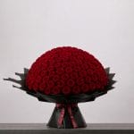A large bouquet of deep red roses arranged in a full, rounded shape, wrapped in elegant black paper and finished with a burgundy ribbon.