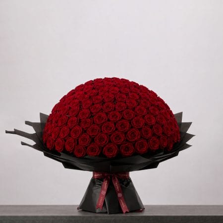 A large bouquet of deep red roses arranged in a full, rounded shape, wrapped in elegant black paper and finished with a burgundy ribbon.