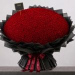 A large bouquet of deep red roses arranged in a full, rounded shape, wrapped in elegant black paper and finished with a burgundy ribbon.