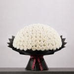 A large bouquet of elegant white roses arranged in a full, rounded shape, wrapped in sophisticated black paper and finished with a refined ribbon.