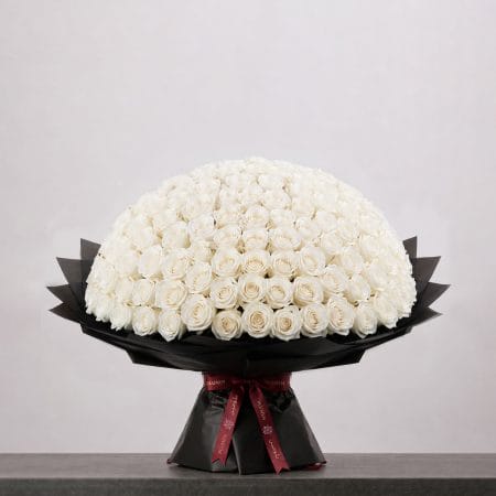 A large bouquet of elegant white roses arranged in a full, rounded shape, wrapped in sophisticated black paper and finished with a refined ribbon.