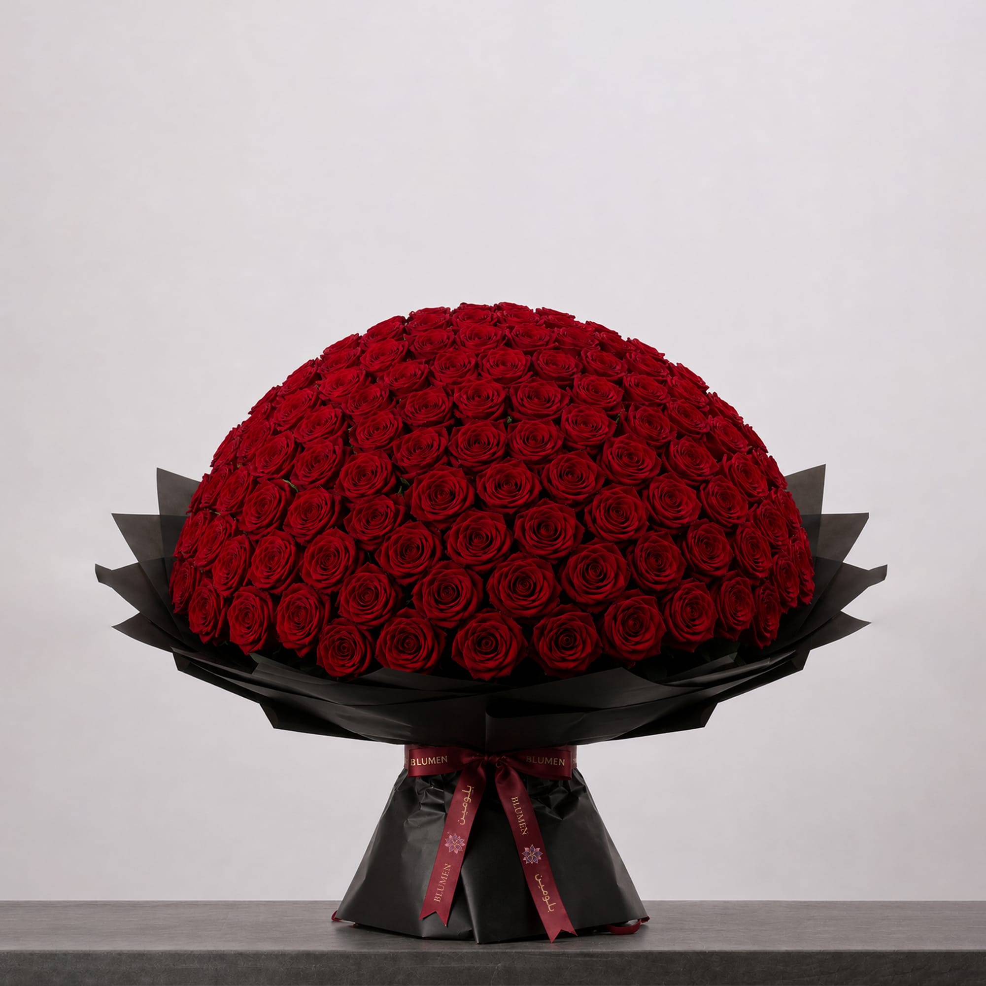 A large bouquet of deep red roses arranged in a full, rounded shape, wrapped in elegant black paper and finished with a burgundy ribbon.