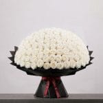 A large bouquet of elegant white roses arranged in a full, rounded shape, wrapped in sophisticated black paper and finished with a refined ribbon.