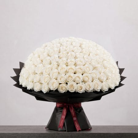 A large bouquet of elegant white roses arranged in a full, rounded shape, wrapped in sophisticated black paper and finished with a refined ribbon.