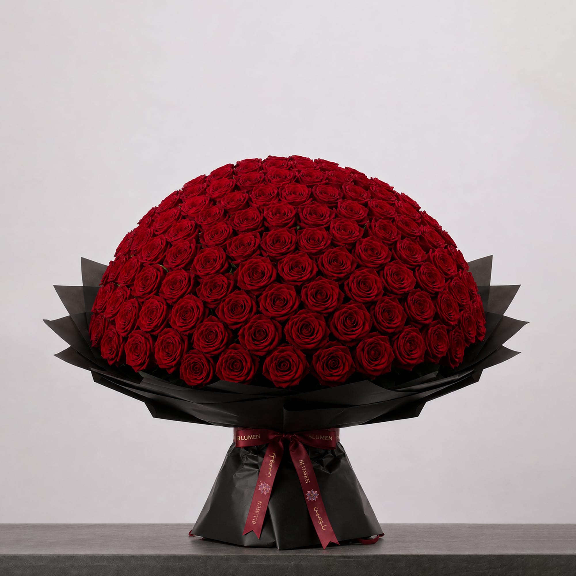 A large bouquet of deep red roses arranged in a full, rounded shape, wrapped in elegant black paper and finished with a burgundy ribbon.